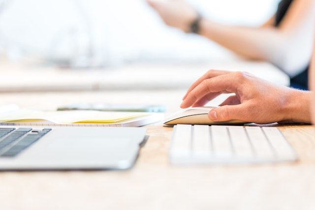 hand on a computer mouse with a wireless keyboard and a laptop on a desk