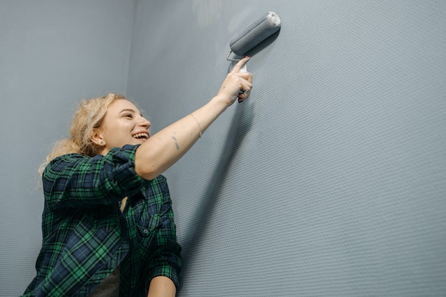 person in a green plaid shirt using a paint roller to paint a wall grey